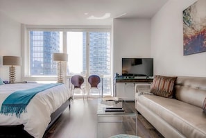 Una habitación de hotel moderna con cama, sofá, escritorio con televisor y un amplio ventanal con vista a la ciudad.
