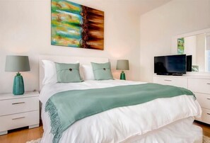 A bedroom with a large bed, two bedside tables, a TV, and a painting on the wall. - Private Fully Furnished 1BR | Washer & Dryer | Midtown West | by GLS (New York)