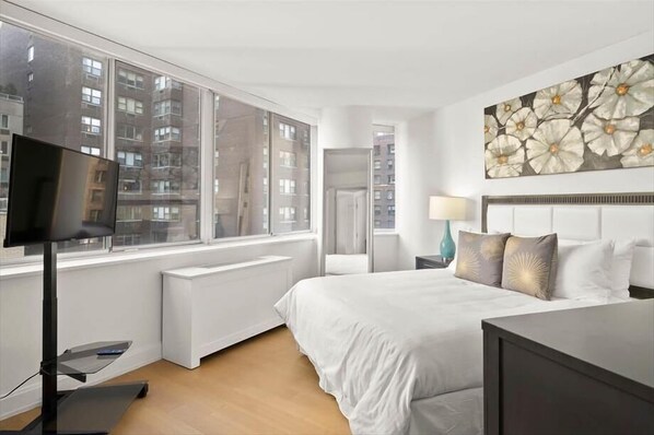 A modern hotel room with a large window, a bed with white bedding, a flat-screen TV, and a wall-mounted artwork.
