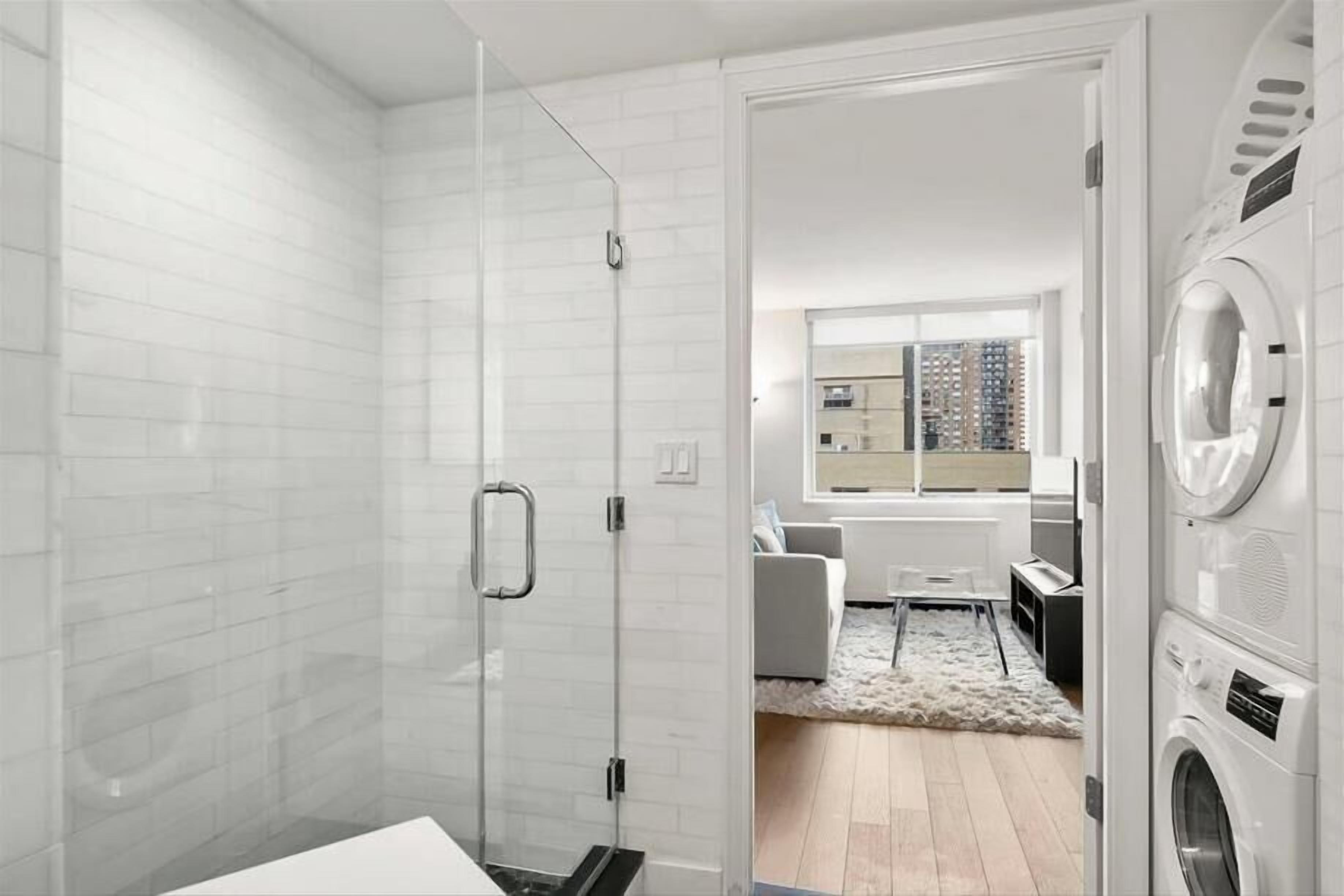 A modern bathroom with a glass shower enclosure, a built-in washing machine, and a view of a cityscape through a large window.