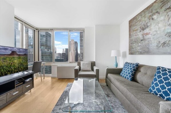 A modern living room with a glass coffee table, grey sofa, and a city view through large windows. - Gorgeous 1BR | Full Kitchen + Gym | Midtown (New York)