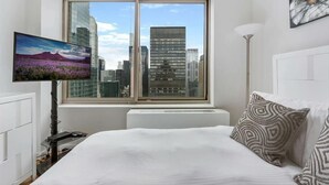 A modern hotel room with a large window offering a city view, a flat-screen TV, and a bed with patterned pillows. - Upscale 2BR | Gym + Terrace | Midtown (New York)