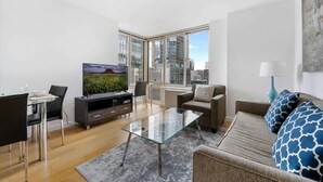 A modern living room with a glass coffee table, a sofa, a TV, and a dining area with chairs and a table. - Upscale 2BR | Gym + Terrace | Midtown (New York)