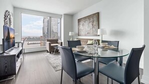 A modern living room with a glass dining table, comfortable seating, a large window with a city view, and a flat-screen TV. - Upper West Side Apartments with Hudson River Views (New York)