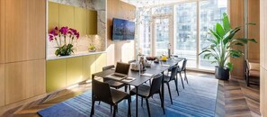 A modern dining area with a long table, chairs, and a laptop. There is a potted plant and a window with a city view.