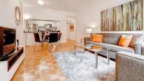 Een moderne woonkamer met een bank, salontafel en televisie. Achtergrond toont een keuken met een eethoek.
