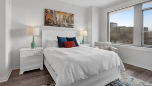 Un dormitorio moderno con una cama grande, mesitas de noche blancas y vistas a la ciudad a través de un amplio ventanal.