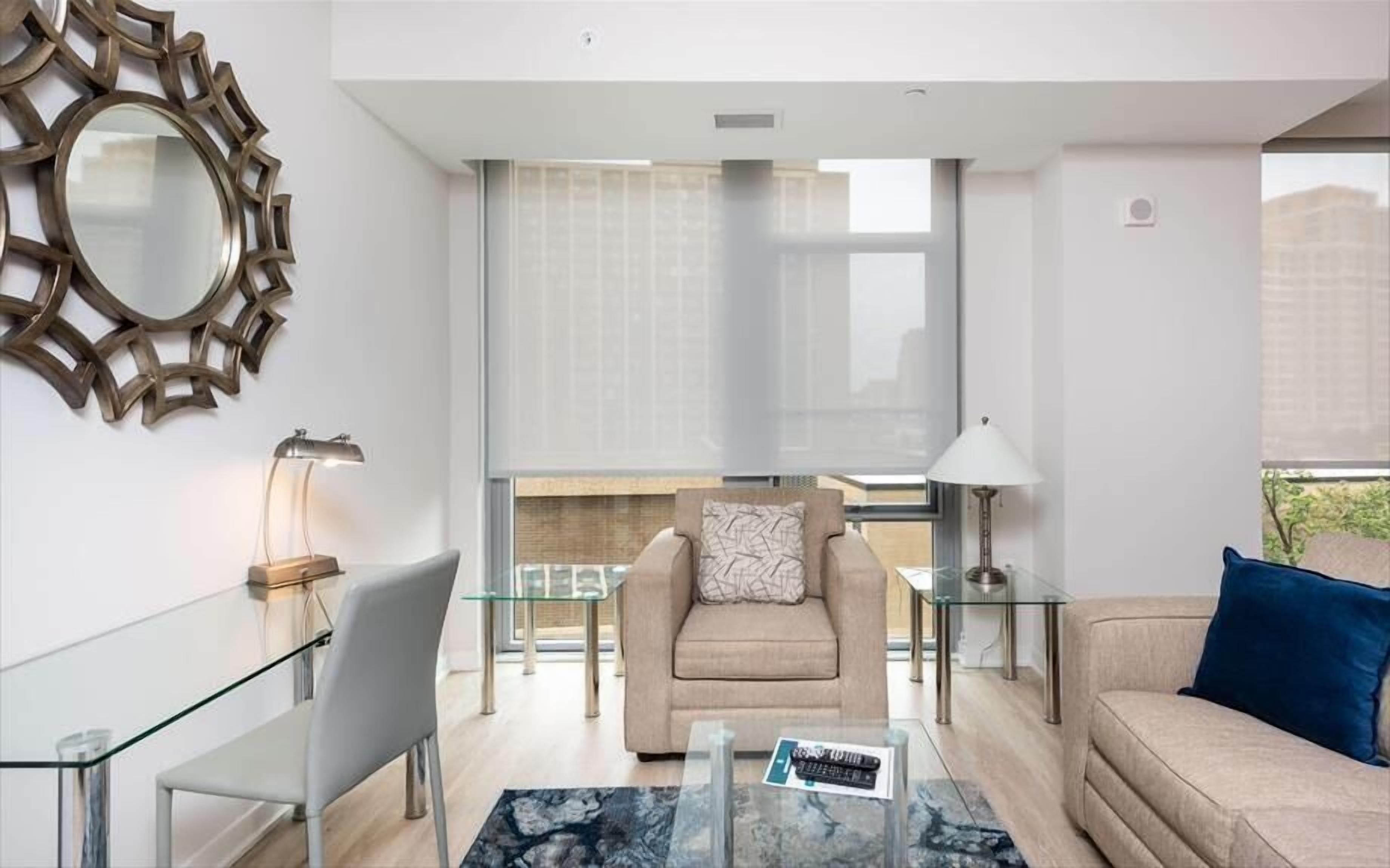 Een moderne woonkamer met een glazen salontafel, een beige bank, een glazen eettafel en een fauteuil met een patroon.