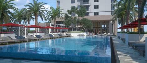 A swimming pool with lounge chairs and umbrellas, surrounded by palm trees and a modern building.