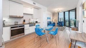 A modern kitchen with white cabinets, stainless steel appliances, and a glass dining table with blue chairs. - Fully Furnished w/ Parking, Pool & Gym | Walk to Metro Mover (Miami)