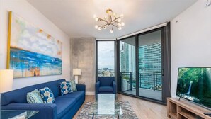 A modern living room with a sofa, a glass coffee table, a TV on a wooden stand, and a large window with a city view. - Fully Furnished w/ Parking, Pool & Gym | Walk to Metro Mover (Miami)
