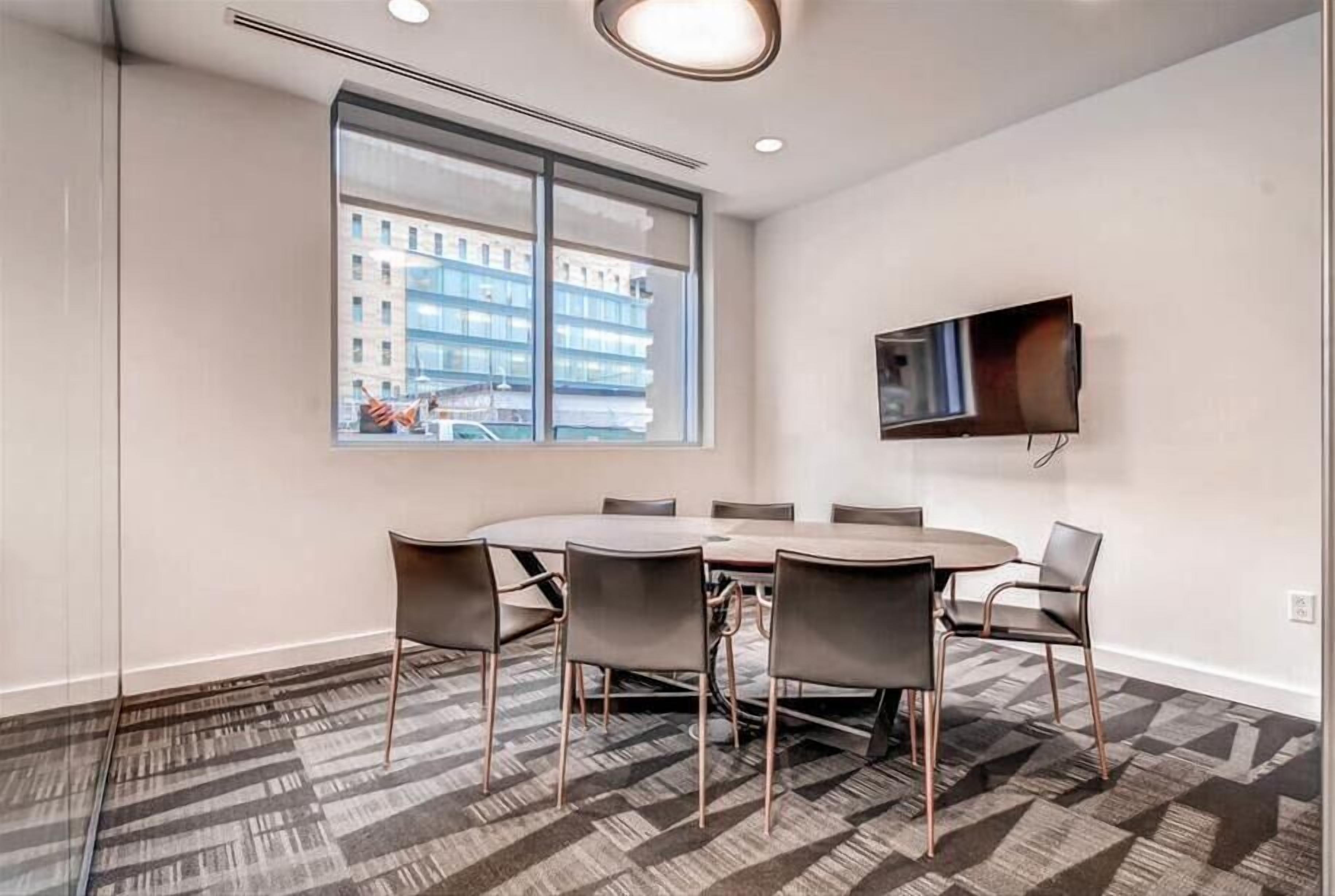 A conference room with a large round table, six chairs, a flat-screen TV mounted on the wall, and a large window offering a view of a building.