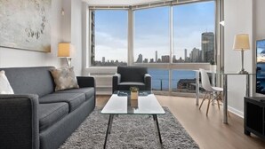 A modern living room with a grey sofa, a glass coffee table, and a view of a city skyline through large windows. - Clean & Spacious 1BR | Work Remote | Free WiFi & Kitchen | Near PATH (Jersey City)