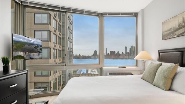 A modern hotel room with a large bed, a TV, a desk, and a view of a cityscape through large windows. - Clean & Spacious 1BR | Work Remote | Free WiFi & Kitchen | Near PATH (Jersey City)
