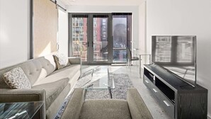 Ein modernes Wohnzimmer mit einer Couch, einem Glastisch, einem Fernseher auf einem Holzschrank und einer Glastür zum Außenbalkon.