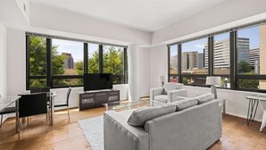 Ein modernes Wohnzimmer mit grauem Sofa, einem Esstisch mit Stühlen, einem Fernseher und großen Fenstern mit Blick auf die Stadt.