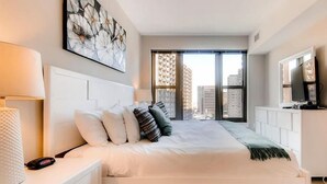 A modern bedroom with a large bed, a TV, and a city view.