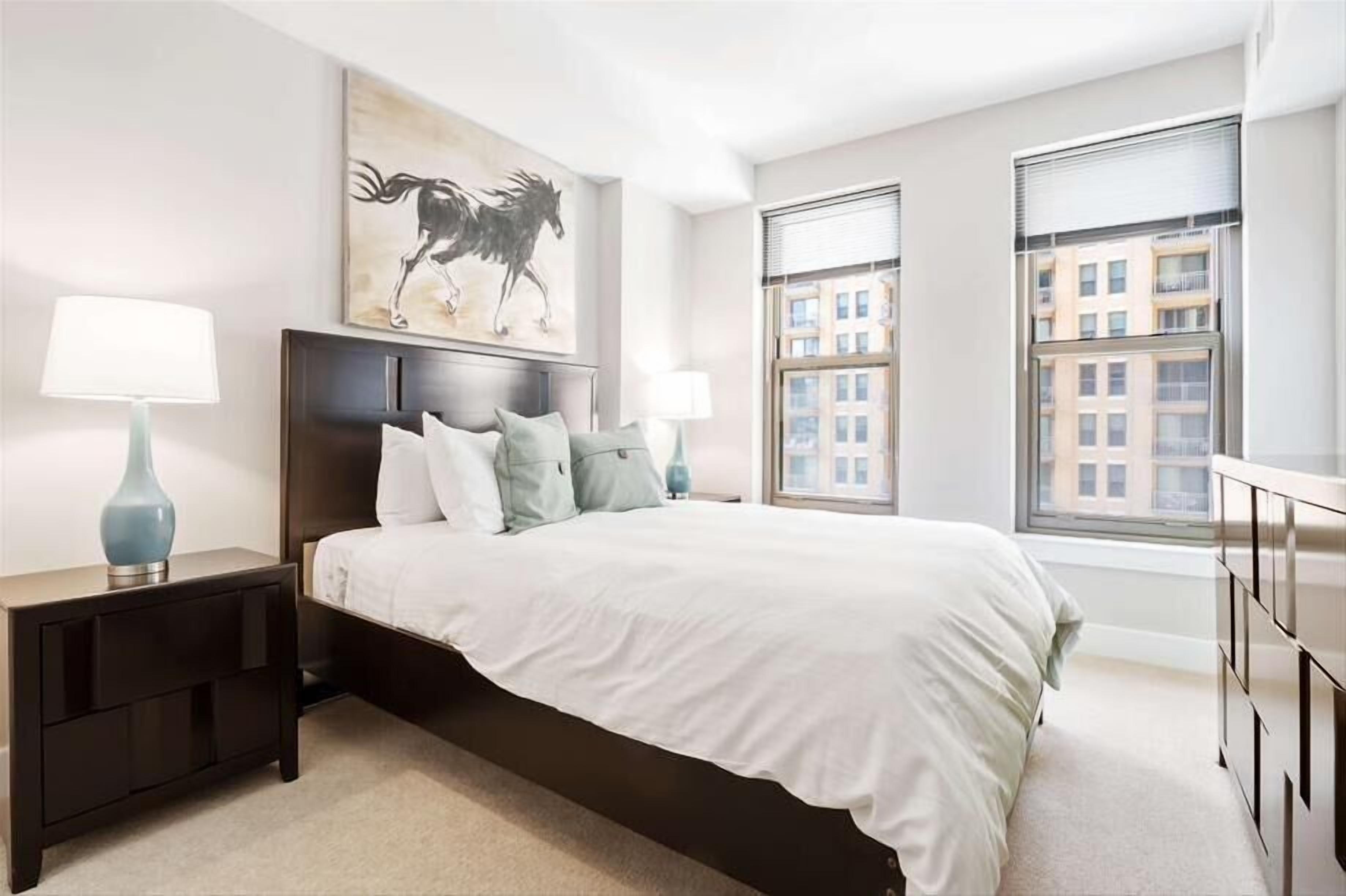 A modern bedroom with a large bed, two bedside tables with lamps, a headboard with a horse painting, and a view of buildings through the window.