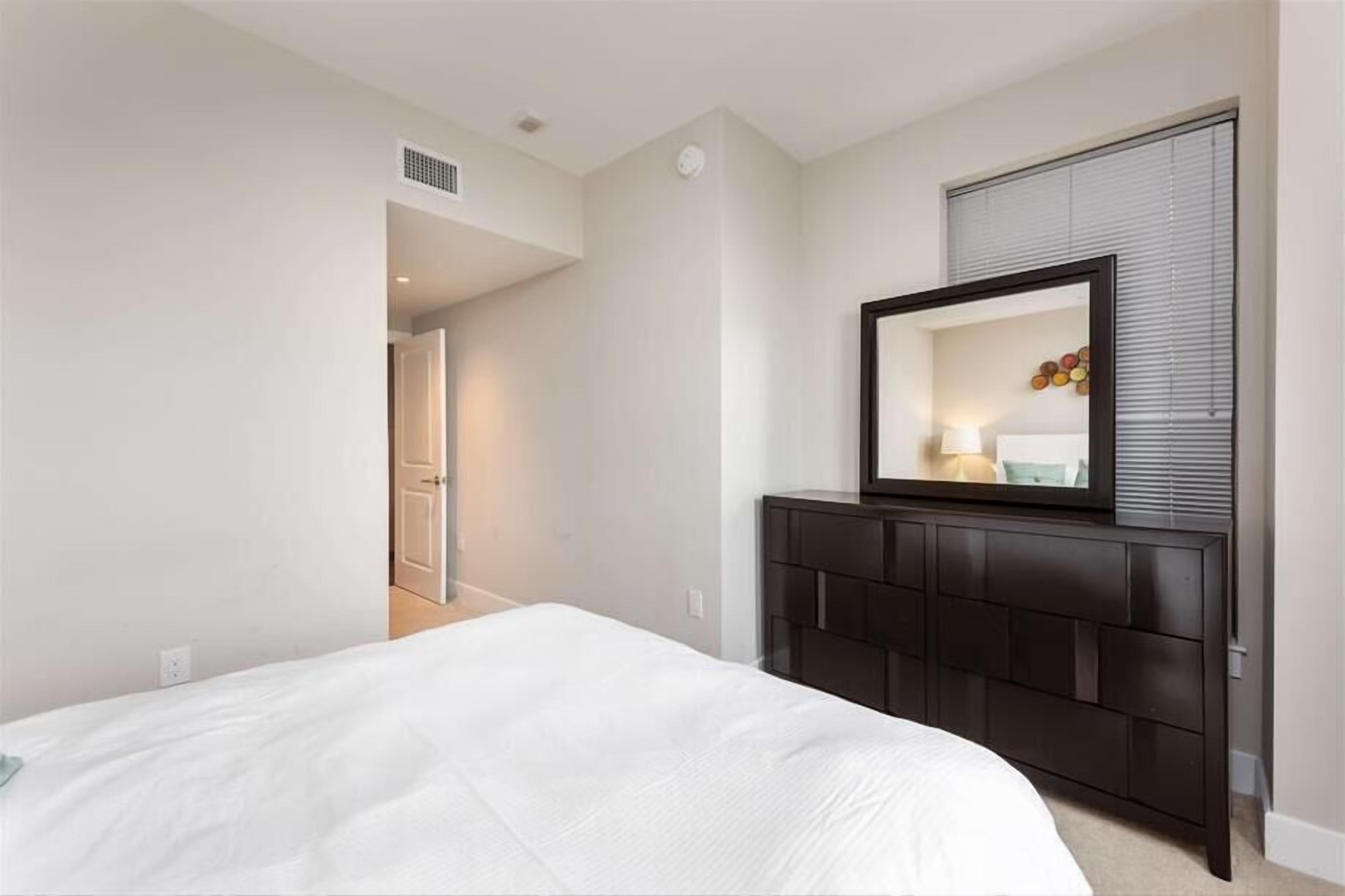 A bedroom with a bed, a dark wooden dresser, a mirror, and a door leading to another room.