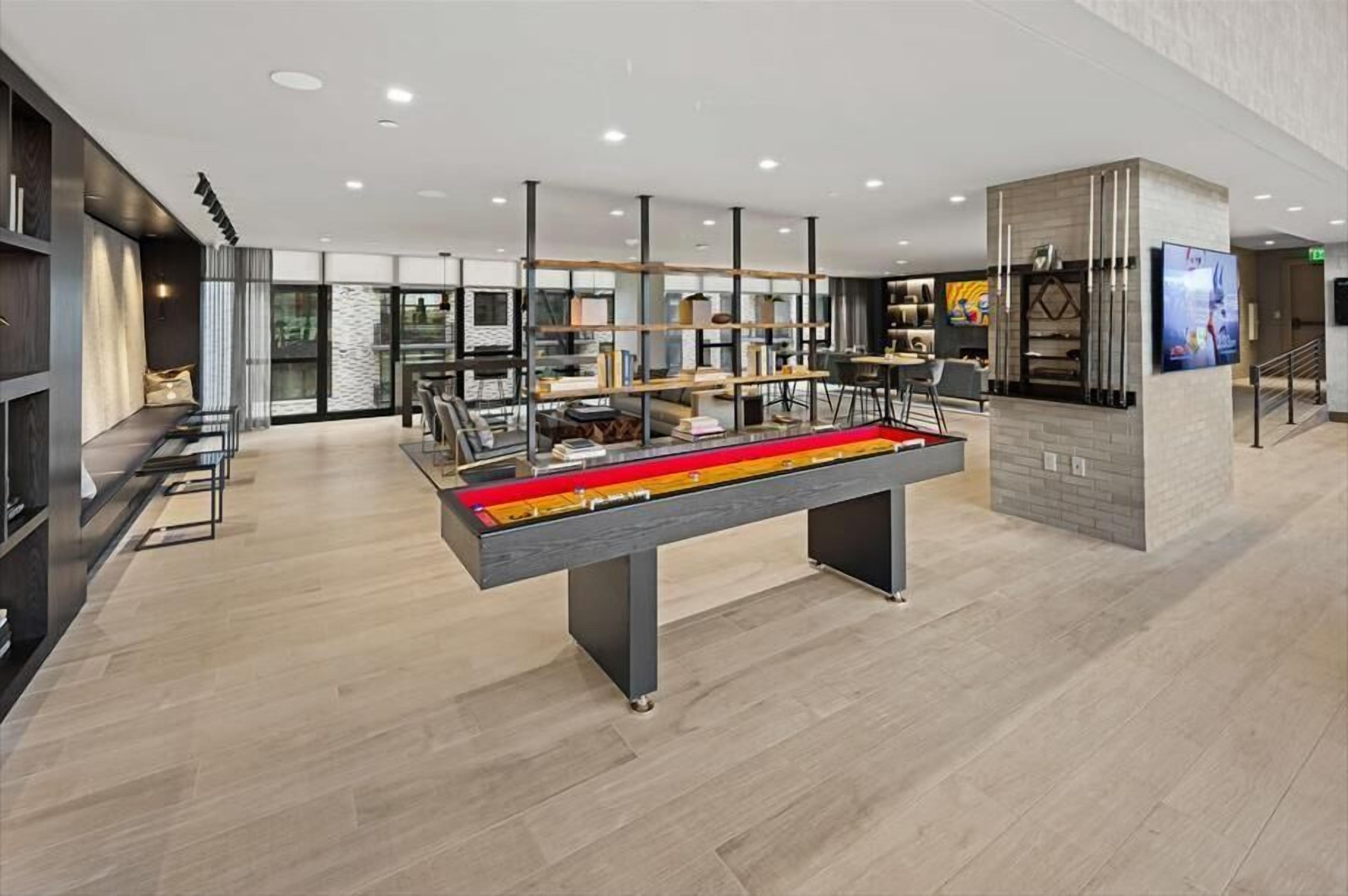 A modern hotel lobby with a central pool table, seating areas, and a glass partition wall.