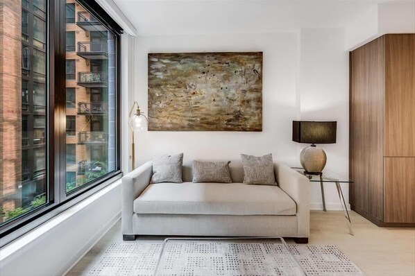 A modern living room with a sofa, a large abstract painting, a lamp, and a view of a building through a large window.
