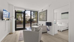 Ein modernes Wohnzimmer mit einer Couch, einem Couchtisch und einem Fernseher. Ein großes Fenster bietet Blick auf Bäume und Gebäude.