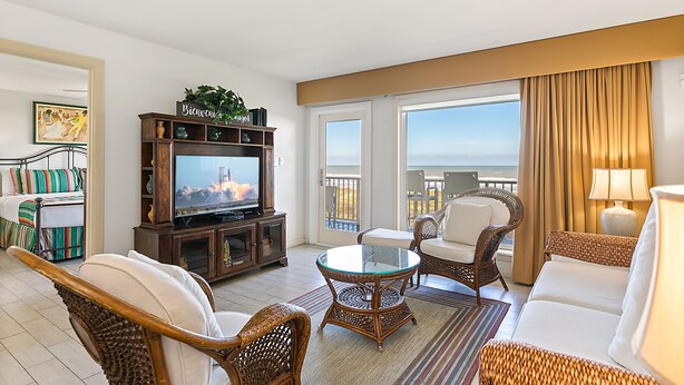 Een ruime woonkamer met een televisie, rieten meubels, een glazen salontafel en een balkon met uitzicht.