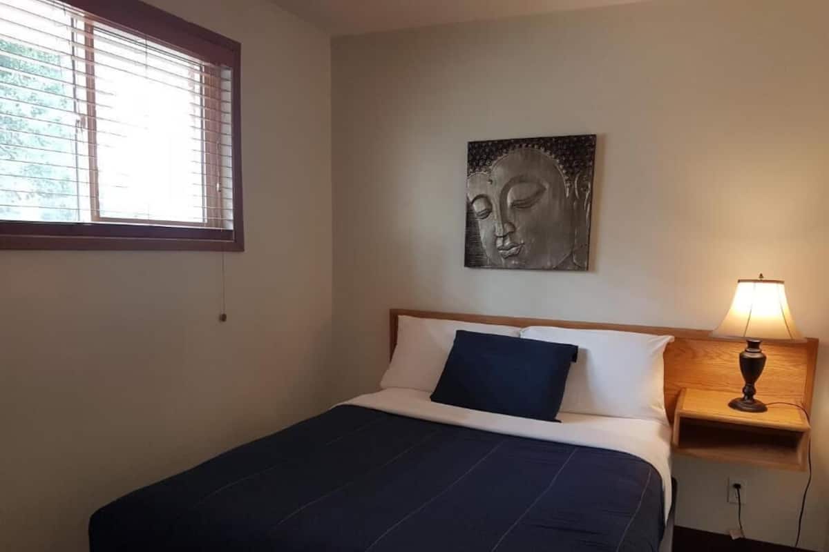 Un dormitorio con cama, mesita de noche con lámpara, ventana con persianas y una obra de arte colgada en la pared.