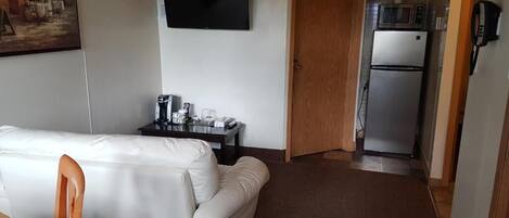 A hotel room with a white sofa, a small table with a lamp, a TV mounted on the wall, and a kitchenette with a fridge and microwave.