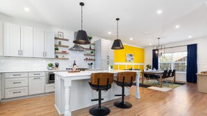 A modern kitchen with white cabinets, a central island with bar stools, pendant lighting, and a dining area with a yellow accent wall. - Hot Tub • Fire Pit • Chef’s Kitchen • Near Uptown (Charlotte)