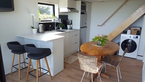 Een kleine keuken met een wit aanrecht, een roestvrijstalen waterkoker, een koffiezetapparaat en een spoelbak. Er staan twee zwarte barkrukken bij een ronde houten tafel met een rieten stoel. In de achtergrond is een wasmachine te zien.