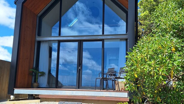 Een modern huis met een grote glazen voorgevel, een houten buitenkant en een klein terrasje met stoelen.