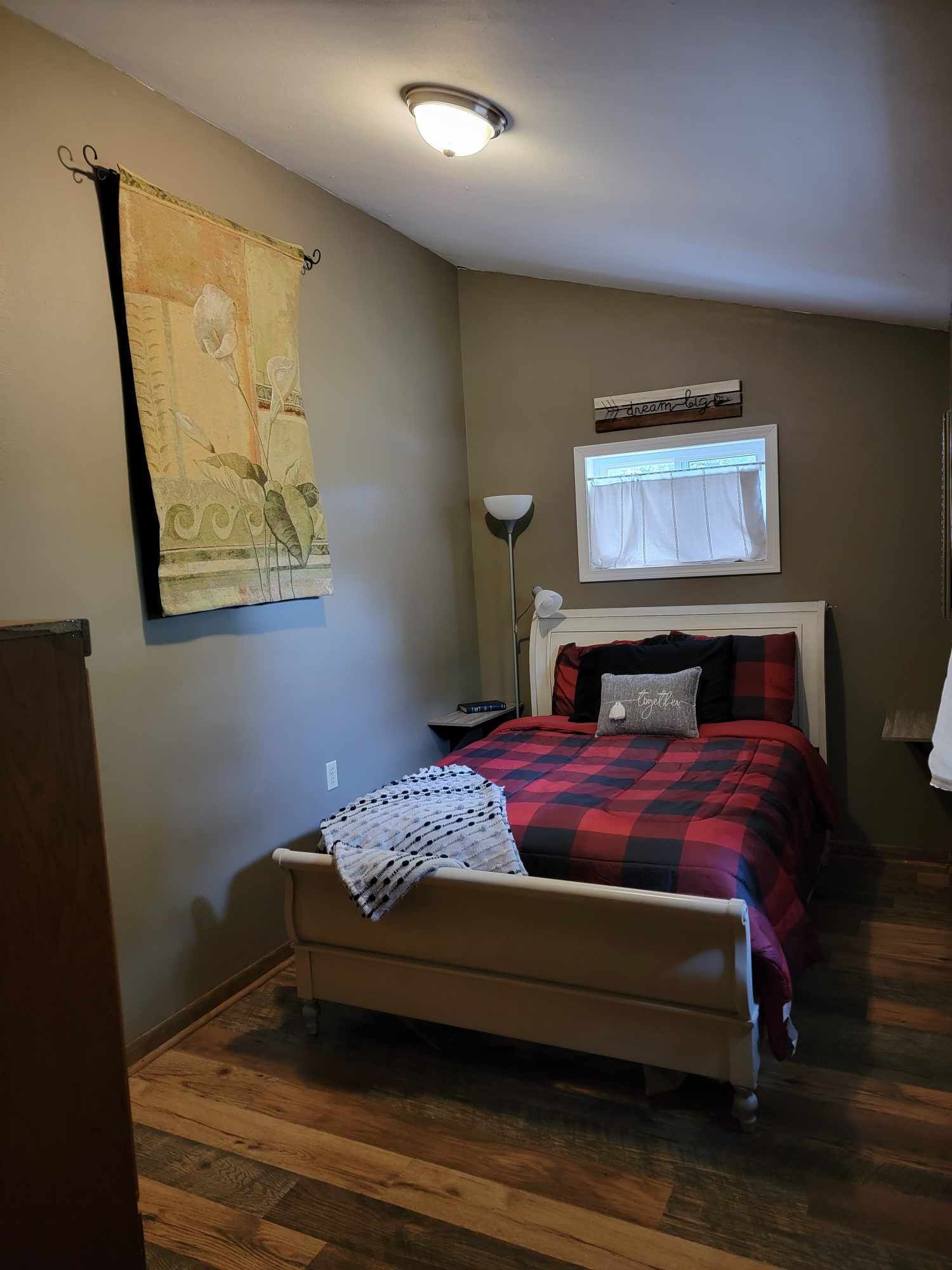 Une chambre à coucher avec un lit, une lampe de chevet, une fenêtre avec des rideaux, une tapisserie au mur et un plancher en bois.