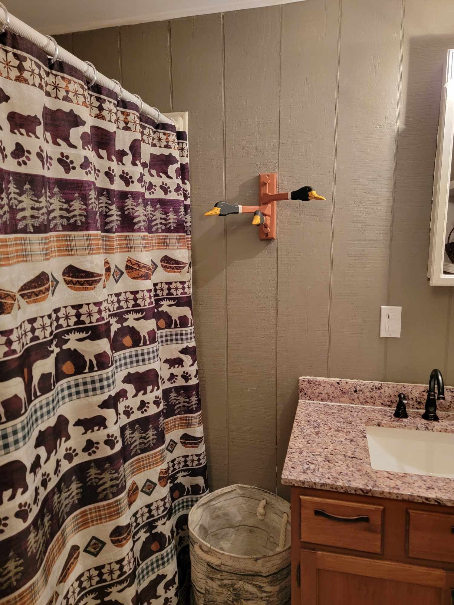 A bathroom with a patterned shower curtain, a wall-mounted bird decoration, a granite countertop sink, and wooden cabinetry.