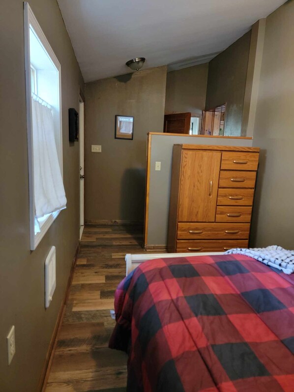 A narrow hallway with a wooden dresser, a bed with a checkered blanket, and a window with a white curtain. - Rustic Cabin on River just off the beaten path (Onaway)
