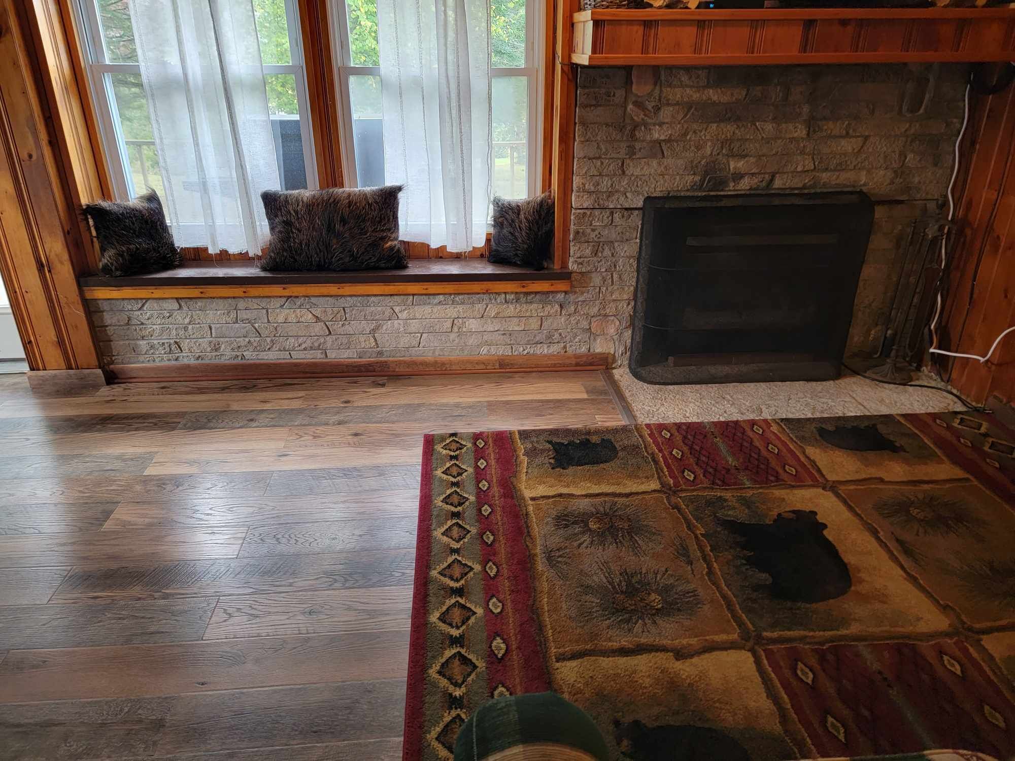 A room with a stone fireplace, a window with curtains, and a patterned rug on a wooden floor.