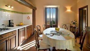 A dining area with a round table set for four, wooden chairs, a kitchen with a stove and coffee maker, and a view through a window. - Beautiful private villa for 8 guests with A/C, WIFI, private pool, TV, patio and panoramic view (Montelopio)