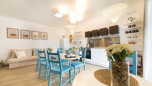 A modern kitchen with white cabinets, a built-in oven, and a stainless steel range hood. There is a dining area with blue chairs and a round table, and a sofa in the background.