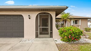 A residential house with a garage door, a front door with a glass panel, and a small garden with a flowering bush.