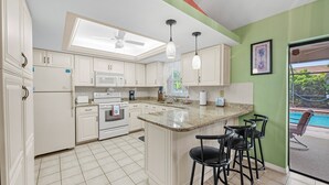 A kitchen with white cabinets, a central island with bar stools, a ceiling fan, and a view of a swimming pool through a glass door.