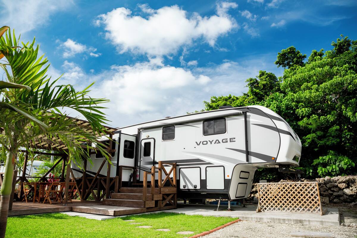 Een witte camper met de tekst 'VOYAGE' erop, geparkeerd in een grasrijk gebied met een houten terras en een trap er naartoe.