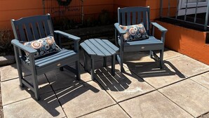 Twee houten stoelen met gestreepte kussens, een tafeltje en een decoratieve krans op een gevel.