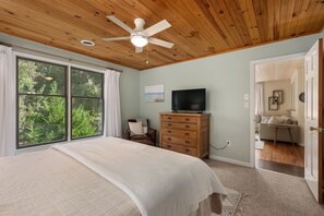 A bedroom with a large bed, a ceiling fan, a TV on a wooden dresser, and a view of greenery through the window.