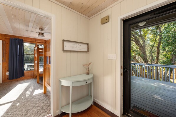 A room with a wooden ceiling, a white wall with a welcome sign, a small white console table, a door leading outside, and a view of trees through the door.