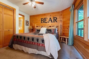 Une chambre confortable avec une tête de lit en bois, un lit recouvert d’une couette à motifs d’ours et une fenêtre donnant sur un paysage verdoyant.