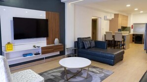 A modern living room with a flat-screen TV, a sofa, a coffee table, and a kitchen area in the background. - Canalily Apt

 (Lekki)