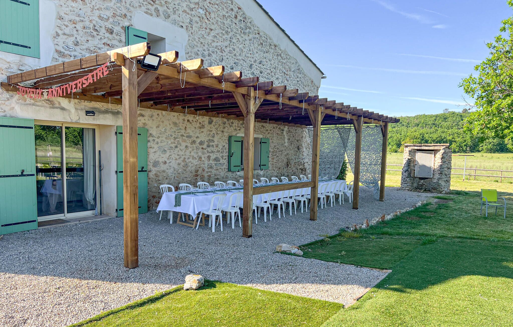 Un espace extérieur abrité, aménagé pour les repas, avec des tables et des chaises, un bâtiment en pierre avec des volets verts, et un chemin de gravier menant à une zone herbeuse.
