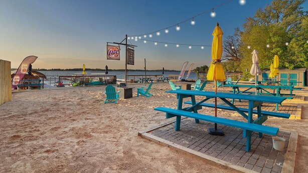 A beachside area with picnic tables and chairs, a flag, and a view of the water. - Chain O` Lakes Beach Penthouse Sunset View and Boat Dock (Ingleside)