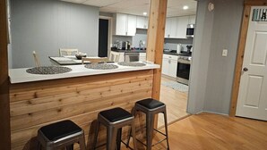 A modern kitchen with wooden accents, a central island with stools, and a view into the dining area. - Chain O` Lakes Beach Penthouse Sunset View and Boat Dock (Ingleside)
