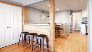 A kitchen with a wooden counter, bar stools, a clock, and a dining area with chairs and a table. - Chain O` Lakes Beach Penthouse Sunset View and Boat Dock (Ingleside)